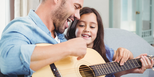 Menina brinca de tocar violão com o pái e Colégio Talento mostra como divertir as crianças em casa nas férias.