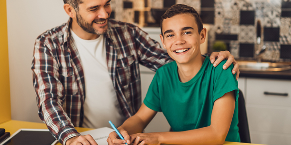 pai com a mão no ombro do filho enquanto ele está sentado fazendo uma tarefa escolar, ambos felizes sorrindo